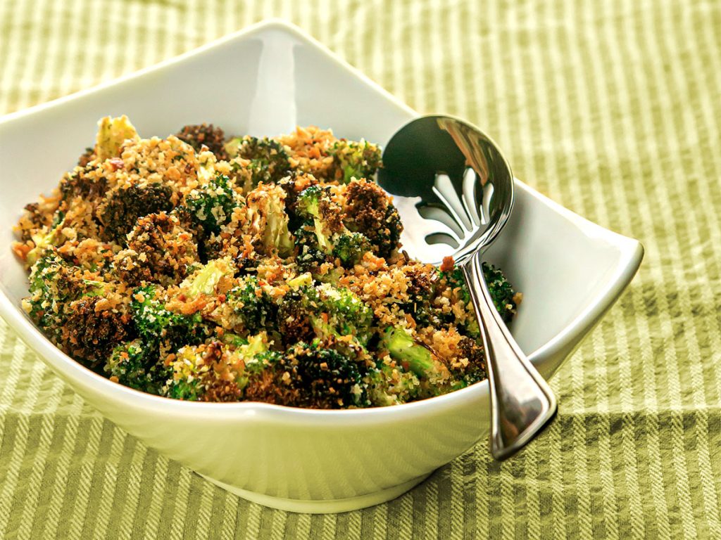A bowl of roasted broccoli topped with toasted Italian breadcrumbs with a spoon on the side