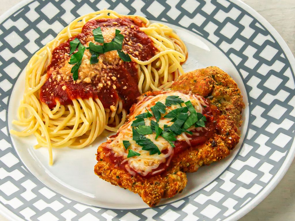 A plate with a serving of spaghetti and chicken parm topped with marinara sauce, cheese, and parsley