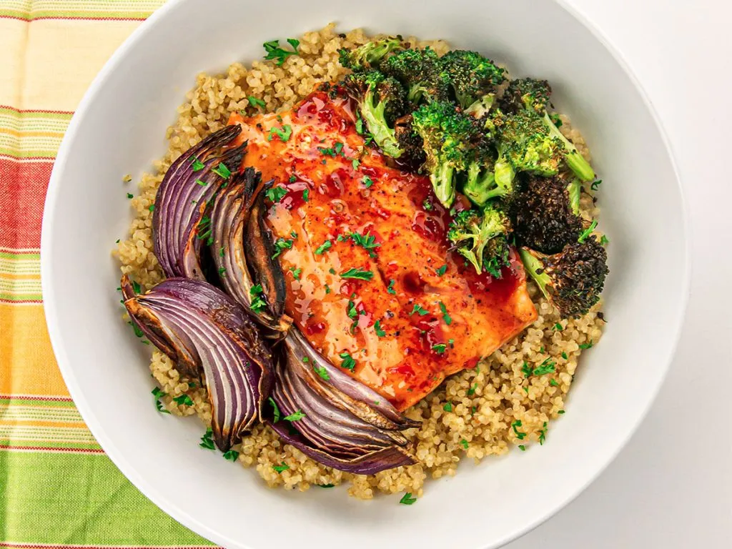 A white bowl with quinoa topped with salmon covered in a sweet chili sauce glaze with roasted broccoli and red onion wedges