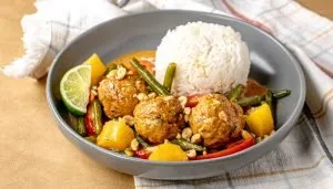 Thai curry meatballs with green beans, pineapple chunks, peanuts, and white rice served in a gray bowl with a lime wedge.
