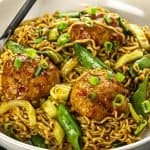 Noodles with meatballs, snap peas, cabbage, and green onions in a white bowl with a fork.