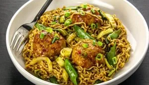 Noodles with meatballs, snap peas, cabbage, and green onions in a white bowl with a fork.