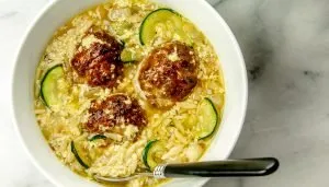 Bowl of soup with meatballs, zucchini slices, egg drop, and noodles, with a black-handled spoon resting inside the bowl on a marble surface.