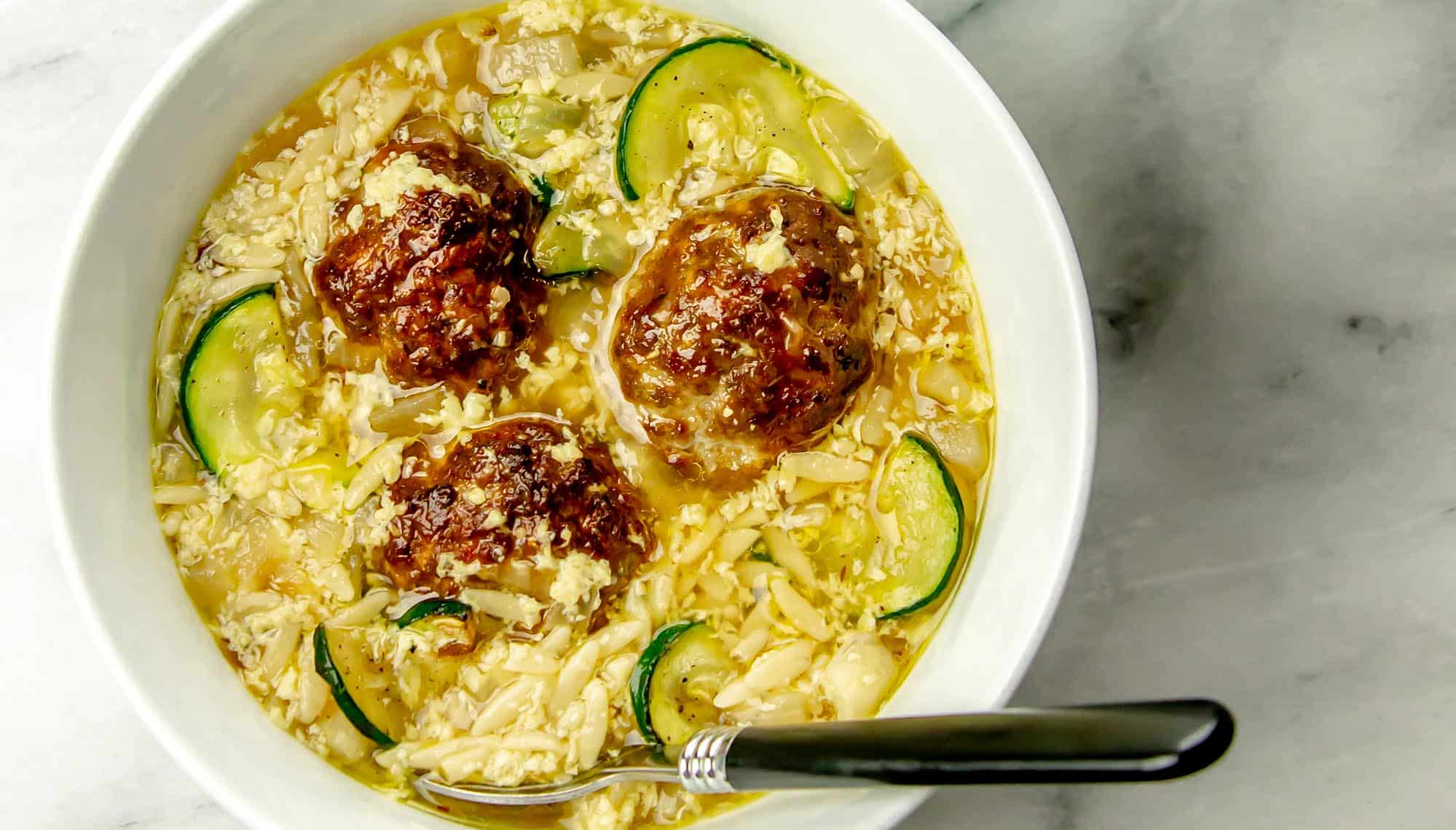 Bowl of soup with meatballs, zucchini slices, egg drop, and noodles, with a black-handled spoon resting inside the bowl on a marble surface.