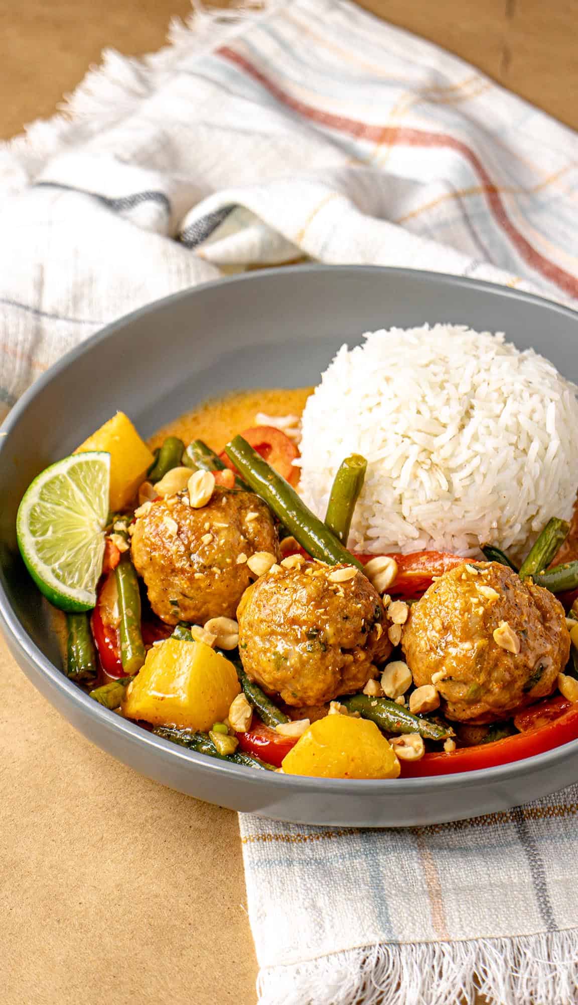 Thai curry with meatballs, pineapple chunks, green beans, peanuts, lime wedge, and a dome of steamed white rice in a gray bowl.