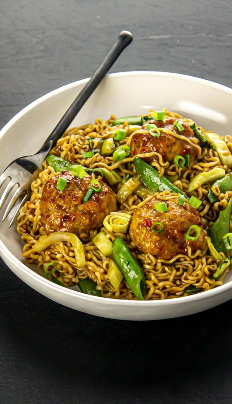 Bowl of noodles with snap peas, chopped green onions, cabbage, and three glazed meatballs, with a fork resting on the bowl rim.