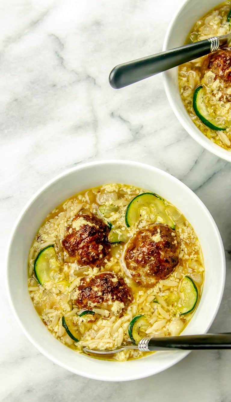 Two bowls of soup with meatballs, orzo, zucchini slices, and egg drop on a white marble surface, each with a black-handled spoon.