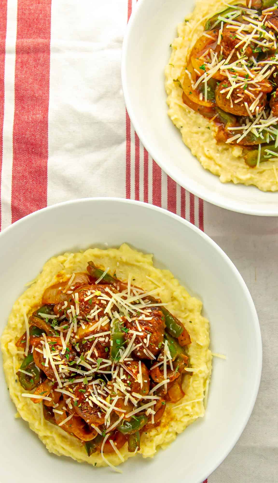 Two white bowls with creamy grits topped with sausage, green bell peppers, onions, tomato sauce, and shredded cheese on a red-striped cloth.