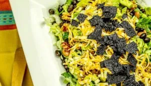 Lettuce salad topped with shredded cheese, black beans, cooked rice, avocado, and blue corn tortilla chip pieces.