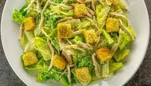 Caesar salad with romaine lettuce, shredded chicken, seasoned croutons, and grated Parmesan cheese on a white plate.