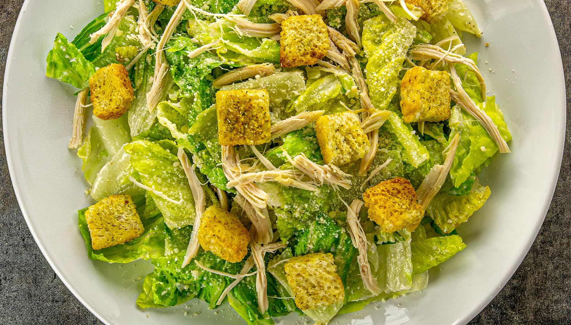 Caesar salad with romaine lettuce, shredded chicken, seasoned croutons, and grated Parmesan cheese on a white plate.