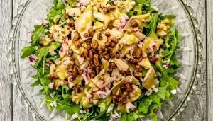 Arugula salad with chopped radishes, chicken pieces, candied nuts, and creamy mustard dressing in a clear glass bowl.