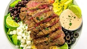 Sliced medium-rare seasoned steak over salad with black beans, corn, avocado, black olives, lime wedges, shredded cheese, and creamy dressing.