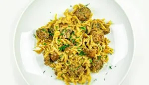 Creamy egg noodles with beef meatballs and mushrooms, garnished with chopped fresh herbs on a white plate.