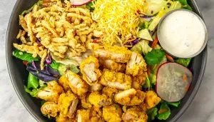 Salad with fried chicken pieces, shredded cheddar cheese, crispy fried onions, lettuce, radish, and a side of ranch dressing in a black bowl.