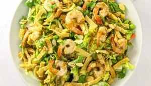 Shrimp salad with lettuce, sliced almonds, sesame seeds, crispy tortilla strips, edamame, and fresh herbs in a white bowl.