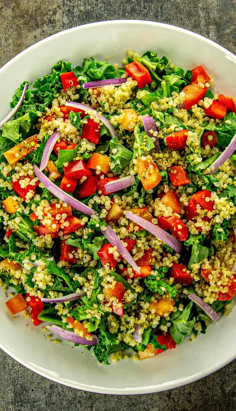 Kale salad with quinoa, red bell peppers, orange bell peppers, and red onion slices in a white bowl on a gray surface.