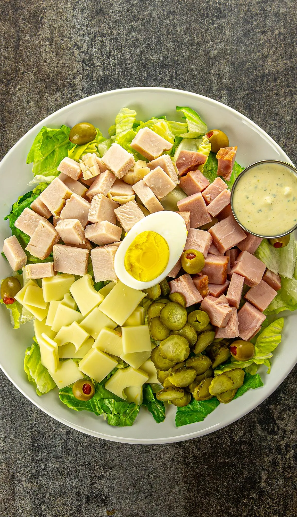 Chopped salad with ham, turkey, cheese, pickles, olives, lettuce, hard-boiled egg half, and a small bowl of dressing on a white plate.