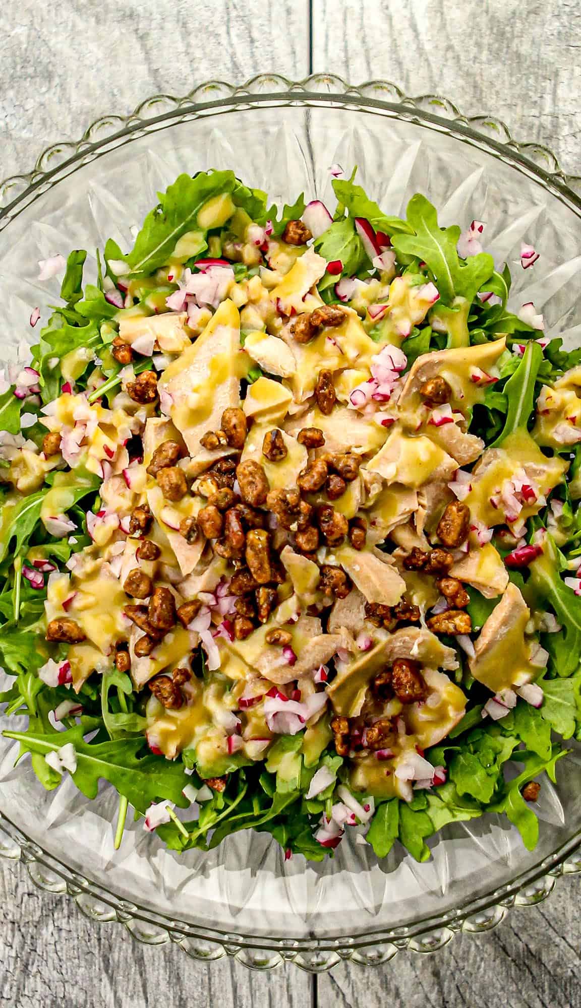 Arugula salad with chopped radishes, shredded chicken, candied nuts, and mustard dressing in a clear glass bowl on wooden surface.