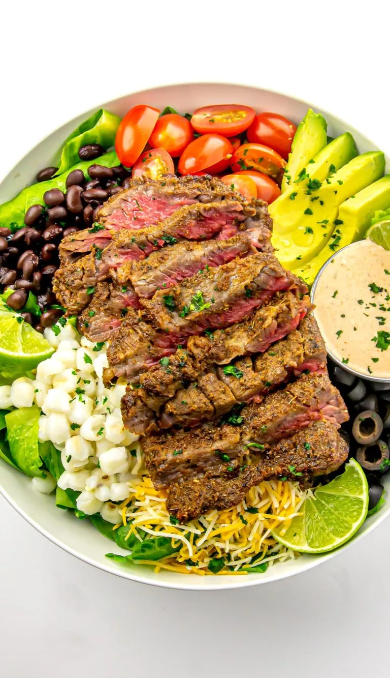 Bowl with sliced medium-rare steak, avocado slices, cherry tomatoes, black beans, corn, shredded cheese, lime wedges, olives, lettuce, and creamy dressing.