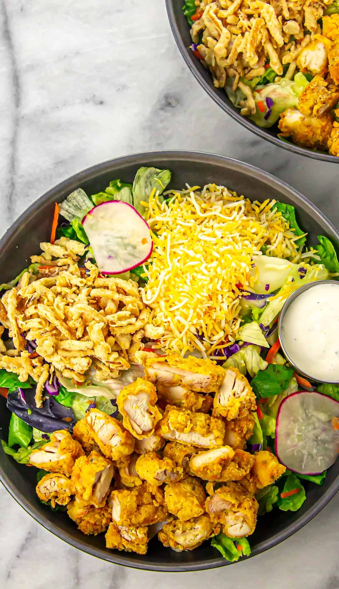 Salad bowl with fried chicken bites, shredded cheese, crispy fried onions, lettuce, radish slices, and a side of ranch dressing.