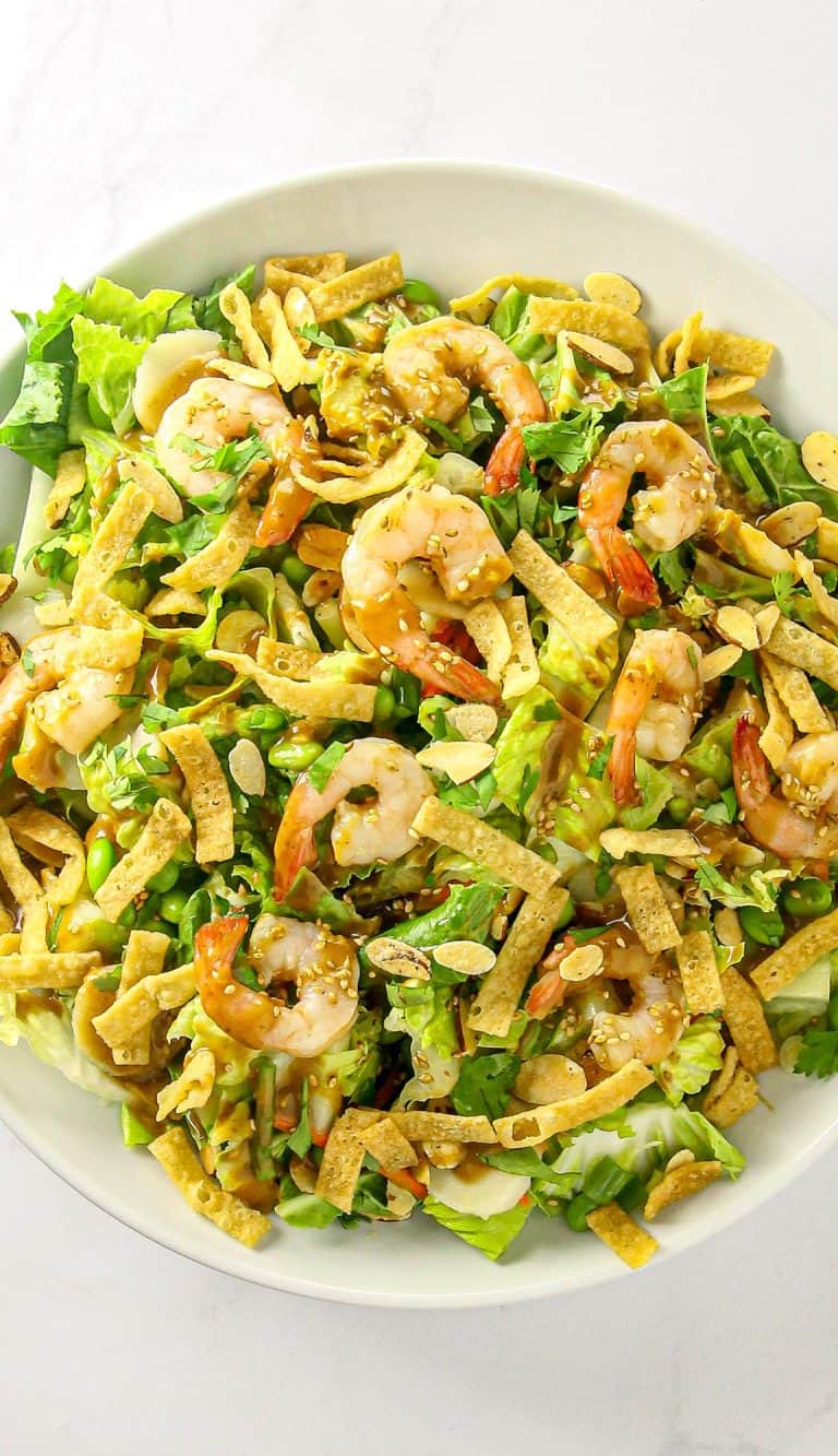 Shrimp salad with lettuce, sliced almonds, crispy tortilla strips, edamame, cilantro, and sesame seeds with dressing on top in a white bowl.