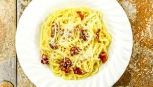Spaghetti carbonara with grated cheese and crispy pancetta on a white plate over a stone textured surface.