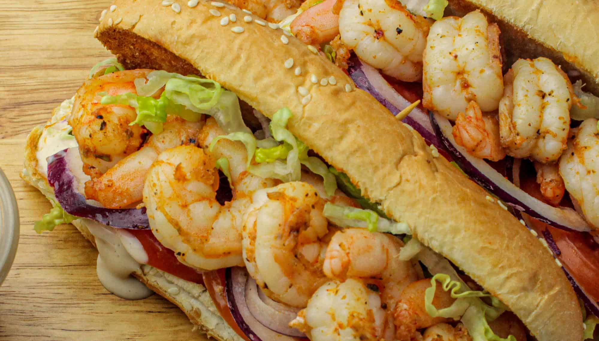 Grilled shrimp sandwich with lettuce, tomato, red onion, and creamy sauce on a sesame hoagie roll on a wooden board.