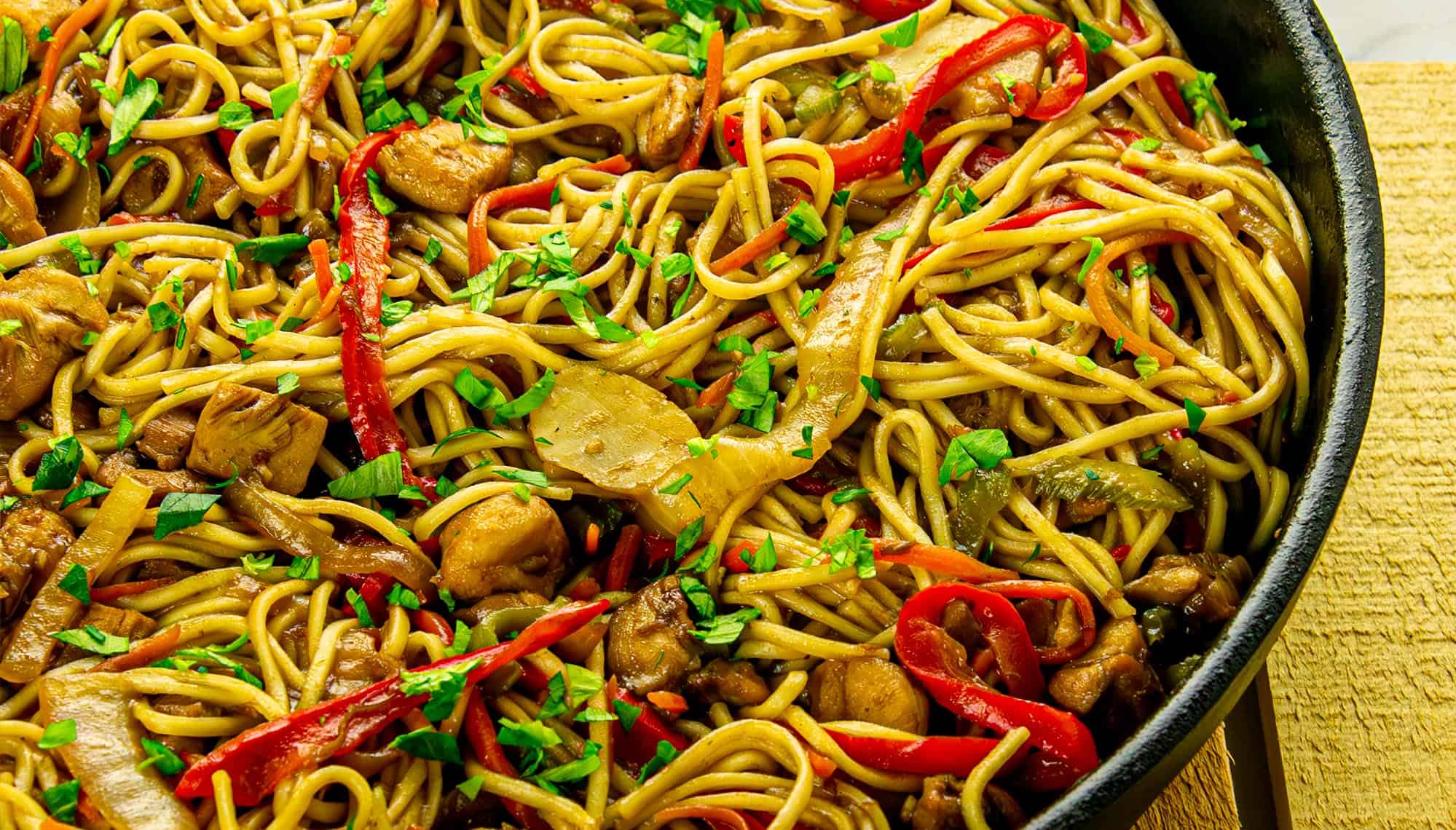 Stir-fried noodles with chicken, red bell peppers, onions, and chopped parsley in a black skillet on a yellow towel.