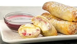 Golden brown dessert egg rolls filled with cream cheese and raspberry, dusted with powdered sugar, served with a bowl of raspberry sauce on a white plate.