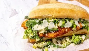 Breaded chicken sandwich with chopped romaine lettuce and shaved Parmesan on a hoagie roll, on white parchment paper.
