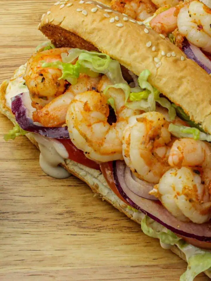 Spicy grilled shrimp sandwich with lettuce, tomato, red onion, and sauce on a sesame hoagie roll, on a wooden surface.