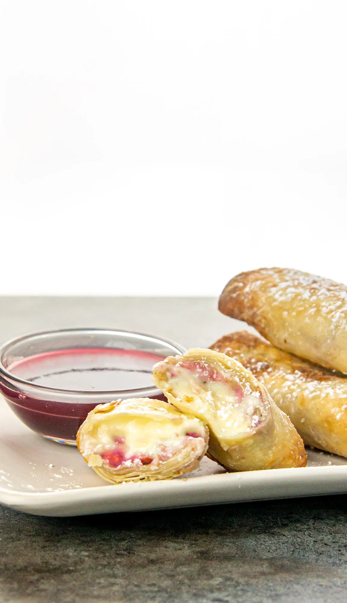 Stack of powdered sugar dusted dessert egg rolls on a plate with one cut in half showing creamy and raspberry filling, next to a bowl of raspberry sauce.