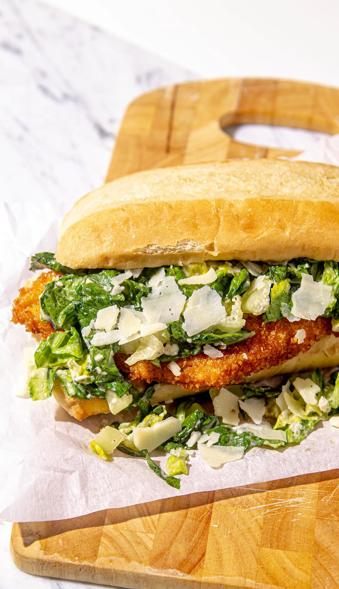 Breaded chicken sandwich with chopped romaine lettuce and shaved Parmesan on a sandwich roll, on white paper and wooden cutting board.