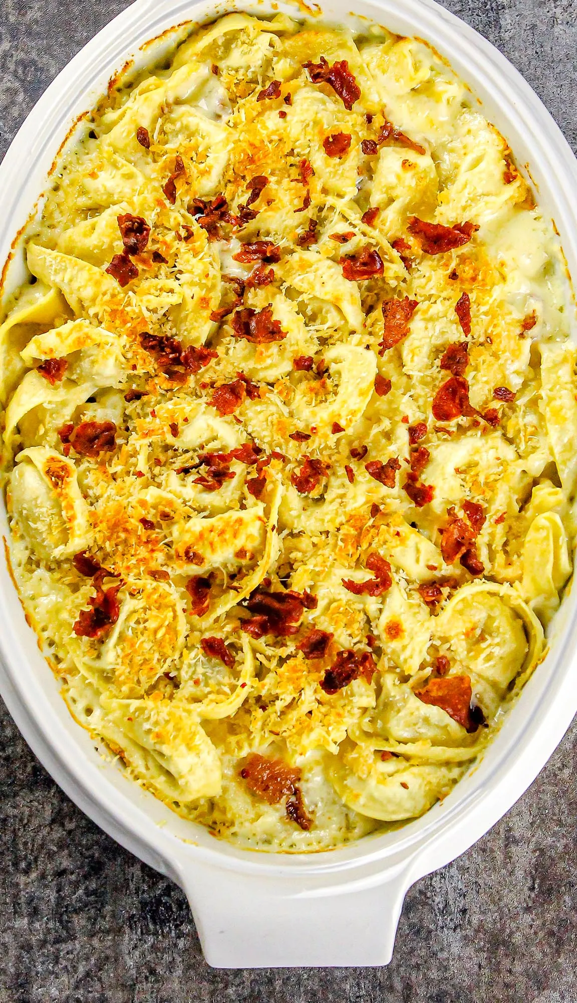 Baked tortellini in creamy sauce, topped with melted cheese and crispy bacon bits in white oval casserole dish.