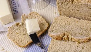 Slices of whole grain bread on a tablecloth, one with a butter pat on a knife and a partially opened butter stick nearby.