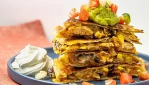 Stacked chicken quesadilla slices with black beans, corn, and melted cheese, topped with guacamole and diced tomatoes, served with sour cream on a blue plate.