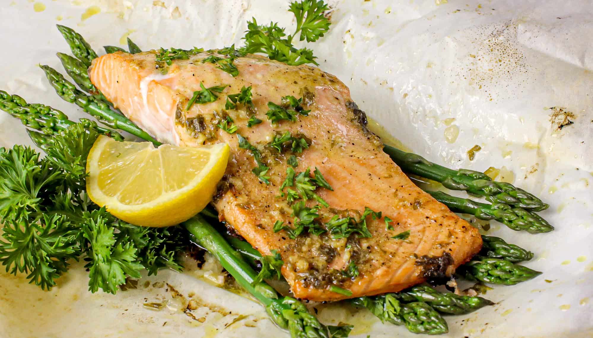Salmon fillet seasoned with herbs on asparagus, garnished with lemon wedge and fresh parsley on parchment paper.