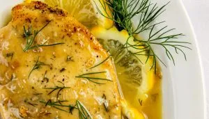 Lemon herb chicken breast garnished with dill, lemon slices, and a lemon sauce on a white plate.