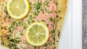 Baked salmon fillet seasoned with herbs and garlic, topped with two lemon slices on parchment paper.