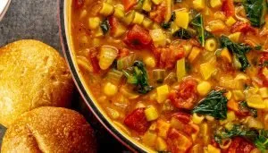 Vegetable soup with tomatoes, greens, chickpeas, and pasta beside two round bread rolls on a dark surface.