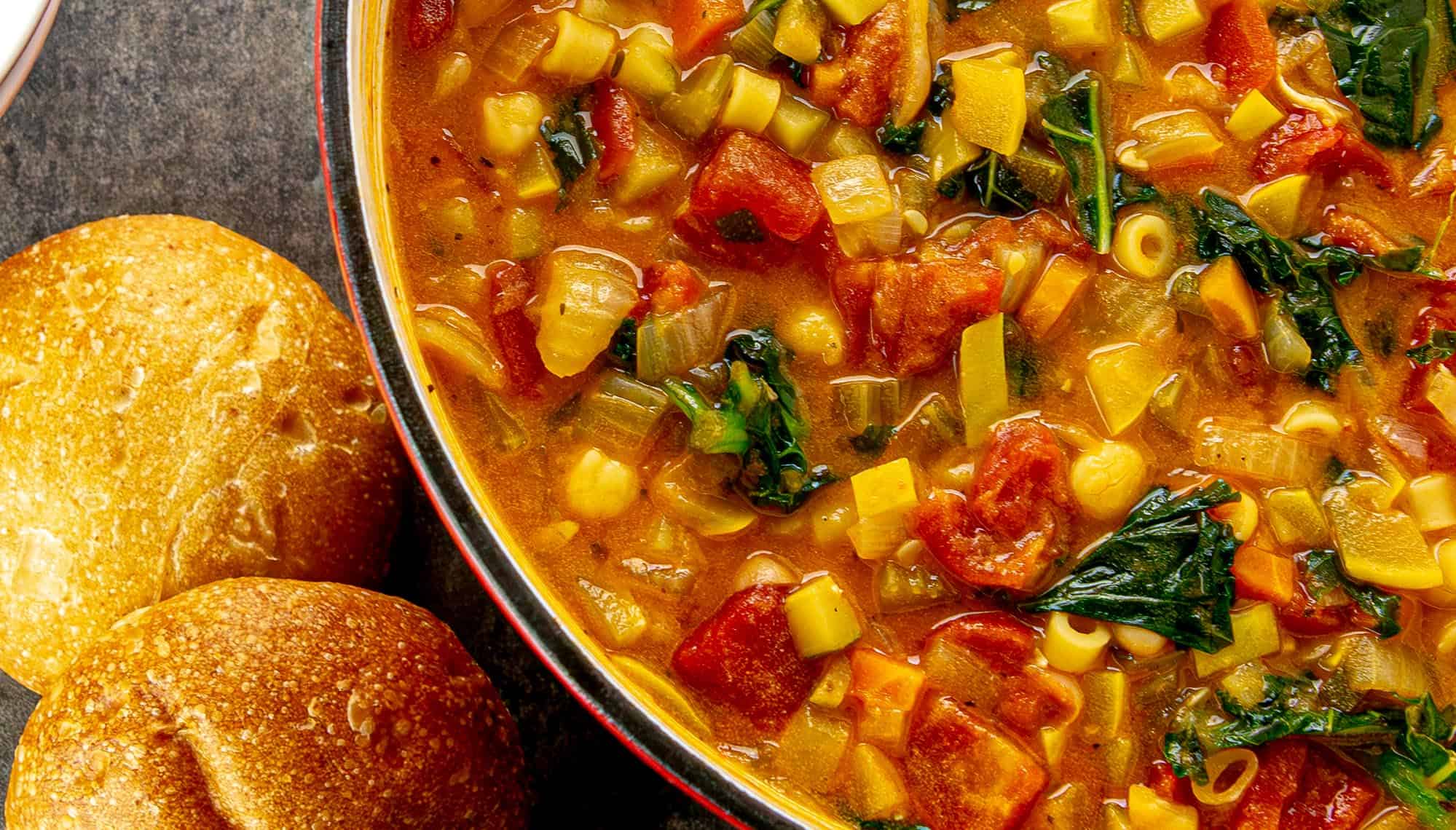 Vegetable soup with tomatoes, greens, chickpeas, and pasta beside two round bread rolls on a dark surface.