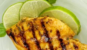 Grilled chicken breast with char marks, garnished with lime slices and mint leaves on a green plate.
