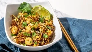 Shrimp Pad Thai noodles garnished with chopped peanuts, green onions, cilantro, and lime wedges in a white bowl with chopsticks on a blue cloth.