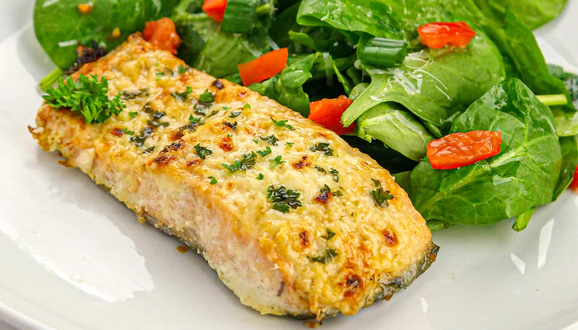 Baked salmon fillet topped with herbs served with fresh spinach salad and chopped tomatoes on a white plate.