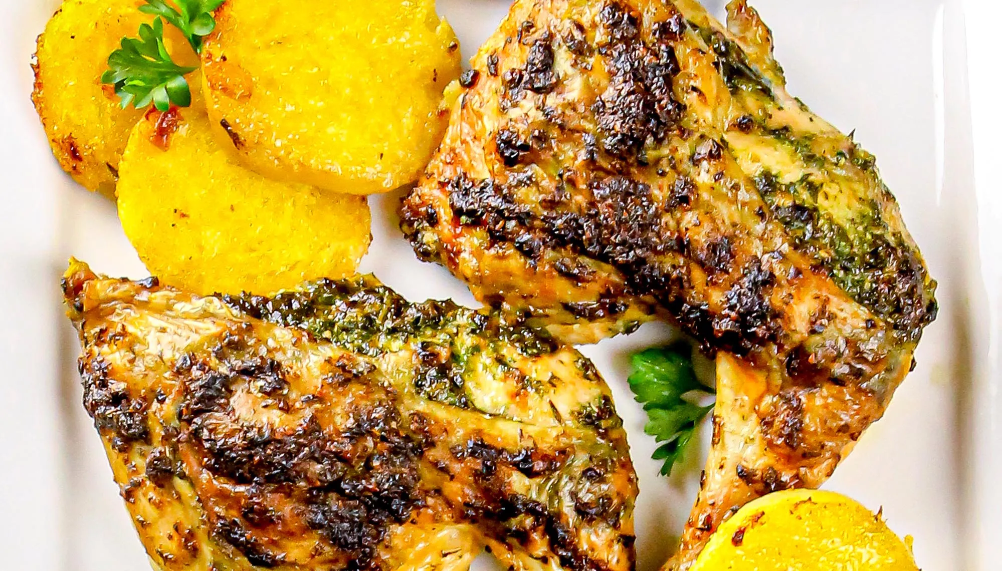 Grilled herb-marinated chicken thighs served with rounds of roasted yellow potatoes and parsley garnish on a white plate.