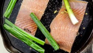 Two raw salmon fillets in a skillet with water, black peppercorns, sliced garlic, and whole green onions.