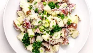 Red potato salad with creamy dressing, chopped green onions, and parsley on a white plate.
