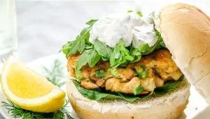 Fish sandwich with lettuce, herb sauce on a bun, lemon wedge, and dill garnish on a white plate