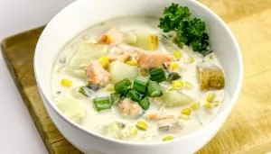 Creamy fish chowder with chunks of potato, corn, green onions, and garnished with parsley in a white bowl on a wooden board.